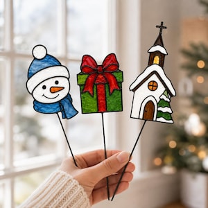 May include: Three stained glass ornaments on thin metal stakes. One is a snowman with a blue hat and scarf, one is a green gift box with a red bow, and one is a white church with a brown door, a cross on top, and a green pine tree beside it.