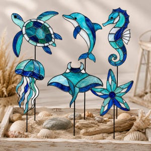 May include: A set of six stained glass sea creature garden stakes featuring a sea turtle, dolphin, seahorse, jellyfish, stingray, and starfish. Each piece is crafted with blue, teal, and white glass panels held together by dark metal solder lines.