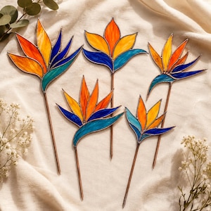May include: Five stained glass bird of paradise flower stakes featuring vibrant orange, yellow, and blue glass petals held together by thin metal solder lines, each attached to a slender copper-colored metal rod for display in soil or pots.