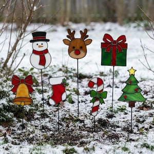 May include: A set of seven stained glass garden stakes featuring holiday icons: a snowman with a red scarf, a reindeer head, a green gift box with a red bow, a yellow bell with a red bow, a red and white stocking, a candy cane, and a green tree.