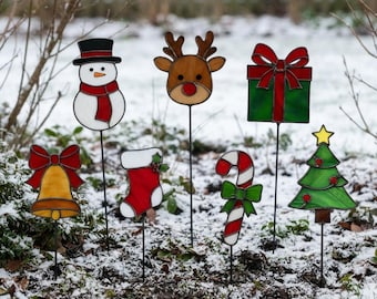 May include: A set of seven stained glass garden stakes featuring holiday icons: a snowman with a red scarf, a reindeer head, a green gift box with a red bow, a yellow bell with a red bow, a red and white stocking, a candy cane, and a green tree.