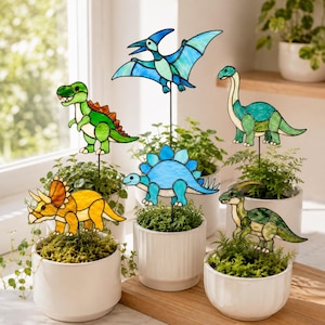 May include: A collection of five stained glass dinosaur plant stakes in various colors and shapes, including a green T-Rex, a blue Pterodactyl, a blue Stegosaurus, a yellow Triceratops, and a green long-necked dinosaur, each on a thin metal rod.