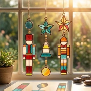 May include: Stained glass window ornaments in the shape of a soldier, star, and Christmas tree. The ornaments are in shades of red, blue, yellow, and green. They hang in front of a window with sunlight streaming through.
