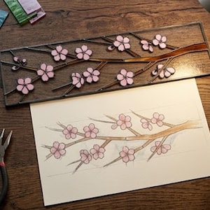 May include: A stained glass panel with a cherry blossom design, featuring pink flowers and brown branches. The panel is on a wooden surface, with a sketch of the design, glass cutting tools, and pieces of coloured glass.