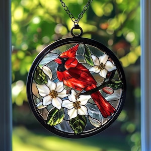 May include: Round stained glass window art featuring a vibrant red cardinal perched on a branch with white flowers and green leaves. The artwork is framed in black and hangs from a chain. The background is a blurred view of green foliage.
