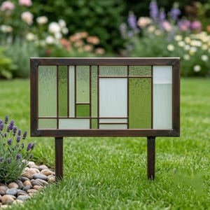 May include: A rectangular garden art piece with a stained glass design. The glass features a geometric pattern of green and white panels within a bronze-colored metal frame. Two metal stakes are attached for ground placement.