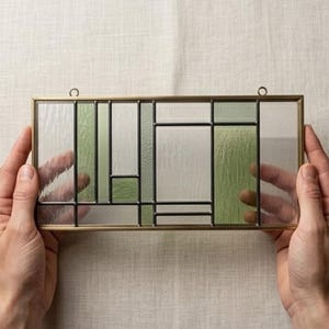 May include: A rectangular stained glass panel with a brass frame. The design features geometric shapes in clear, white, and shades of green. Two small metal loops are attached to the top for hanging. The panel is held by two hands.