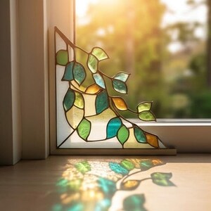 May include: Stained glass corner decoration with a leafy branch design. The leaves are various shades of green, teal, and yellow, framed by a brown border. It sits on a windowsill, with sunlight illuminating the glass.