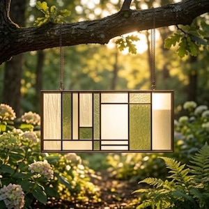 May include: A rectangular stained glass panel with a geometric design, hanging from a chain. The panel features shades of green and cream, framed in a dark metal. The background is a blurred view of a sunlit garden.