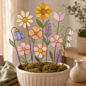 May include: A collection of stained glass flower stakes arranged in a round, ribbed ceramic pot filled with green moss. The set includes various floral shapes in shades of yellow, pink, blue, purple, and white, each with green glass leaves.