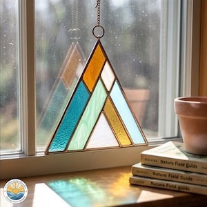 May include: A stained glass suncatcher in a geometric mountain design. The triangular piece features sections of teal, white, gold, and orange glass, framed in a copper-colored metal. It hangs from a chain, reflecting light. The logo of "CraftoraGlassStudio" is visible.