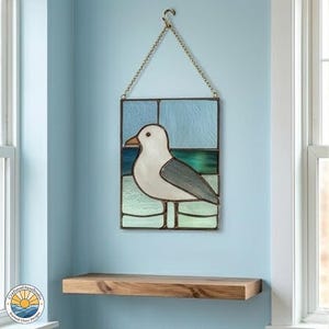 May include: Stained glass panel depicting a seagull. The artwork shows a white seagull with grey wings and a yellow beak, set against a blue and green glass background. It is suspended by a chain and hook.