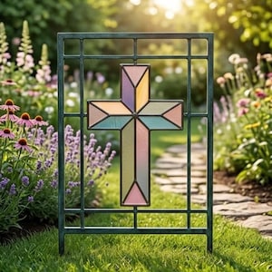 May include: A decorative garden cross with a stained-glass design. The cross features a multi-colored pattern in shades of pink, green, yellow, and purple, set within a dark green metal frame. The cross is placed in a garden setting with green grass and colorful flowers.