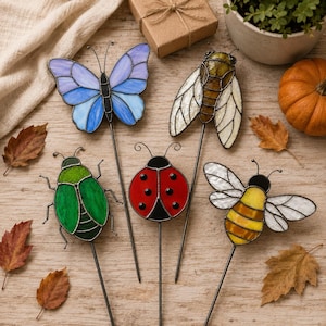 May include: Five stained glass insect garden stakes featuring a blue and purple butterfly, a brown cicada, a green beetle, a red ladybug with black spots, and a yellow and black striped bee, each attached to a thin metal rod for garden display.