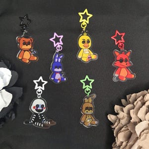 May include: A collection of seven character keychains. Each keychain features a different cartoon character in various colors, including brown, yellow, red, purple, and white. Each character is attached to a star-shaped charm and a metal keyring.
