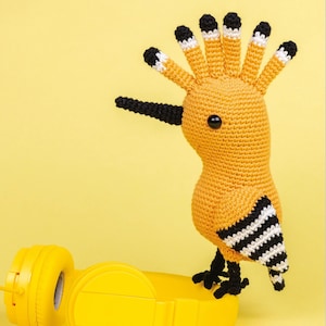 May include: A handcrafted crochet bird in yellow, black, and white. The bird features a long, black beak and a fan of upright feathers. It stands on black feet, with a black and white striped wing. Yellow headphones are in the foreground.
