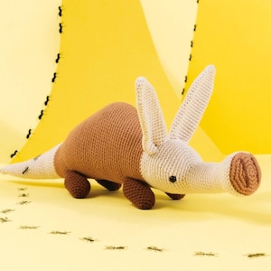 Può includere: Un giocattolo di oritteropo all'uncinetto nei toni del marrone, beige e nero, con lunghe orecchie e un muso caratteristico. Il peluche fatto a mano è ambientato su uno sfondo giallo con una serie di illustrazioni di piccoli insetti.