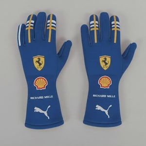 Peut inclure: Une paire de gants de course bleus avec des détails blancs et jaunes. Chaque gant présente un emblème Ferrari jaune, un logo Shell, le texte "RICHARD MILLE" et un logo Puma blanc. Conçus pour la course.