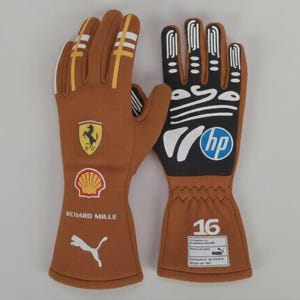 Peut inclure: Une paire de gants de course bruns avec des rayures blanches et jaunes. Les gants présentent les logos Ferrari, Shell et Puma. Le gant gauche porte le texte "RICHARD MILLE" et le gant droit le numéro "16" et le logo HP.