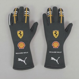 Peut inclure: Une paire de gants gris avec des rayures blanches et dorées sur les doigts. Les gants présentent le logo Ferrari, le logo Shell, le texte Richard Mille et le logo Puma. Les gants sont conçus pour la course.