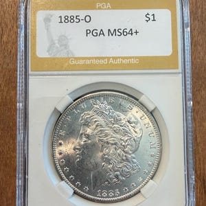 May include: A silver 1885-O Morgan silver dollar coin, graded PGA MS64+, in a clear protective case. The coin features a profile of a woman and the text "$1". The case has a gold label with the text "Guaranteed Authentic".