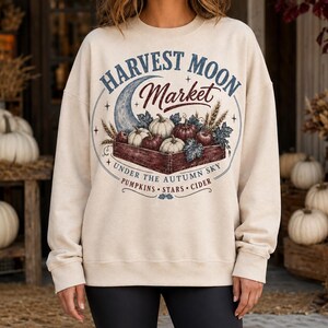 Peut inclure: Un sweat-shirt beige à col rond avec une impression graphique représentant une caisse en bois remplie de citrouilles et de pommes, un croissant de lune et le texte "HARVEST MOON MARKET UNDER THE AUTUMN SKY PUMPKINS STARS CIDER" en bleu et rouge.