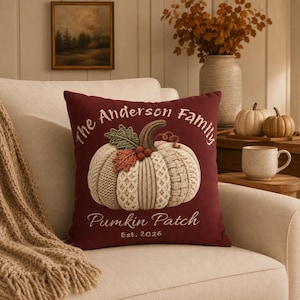 Peut inclure: Un coussin carré bordeaux orné d'un motif central représentant une citrouille crème à la texture tricotée, avec des feuilles vertes et des baies rouges. Le texte indique The Anderson Family, Pumpkin Patch et Est. 2026 en blanc.
