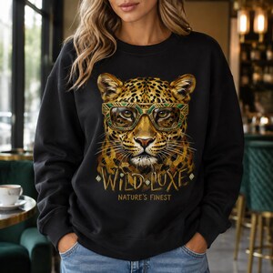 Peut inclure: Un sweat-shirt noir à manches longues avec un imprimé représentant un léopard portant des lunettes à motifs géométriques. Sous le léopard, le texte "WILD LUXE" est écrit en lettres dorées, suivi de "NATURE'S FINEST" en plus petit.