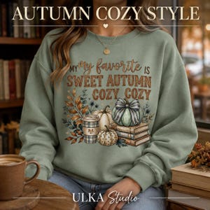 Peut inclure: Un sweat-shirt col rond vert sauge avec le texte "MY FAVORITE IS SWEET AUTUMN COZY COZY" en marron et orange. Le motif comprend une tasse à café à carreaux, trois citrouilles, un nœud rayé et une pile de livres avec des feuilles d'automne.