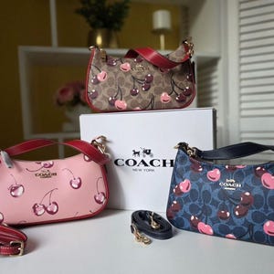 Puede incluir: Tres bolsos Coach con estampados de cerezas. Uno es rosa, otro marrón y otro azul marino. Cada bolso tiene una correa roja y herrajes dorados. El logotipo de Coach es visible en los bolsos y la caja.