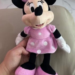 Pode incluir: Uma boneca de pel&uacute;cia da Minnie Mouse, com um vestido rosa com bolinhas brancas, um la&ccedil;o a condizer e orelhas pretas. A boneca tem luvas brancas e sapatos rosa. A boneca tem cerca de 30 cm de altura.