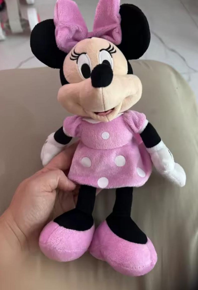 Pode incluir: Uma boneca de pel&uacute;cia da Minnie Mouse, com um vestido rosa com bolinhas brancas, um la&ccedil;o a condizer e orelhas pretas. A boneca tem luvas brancas e sapatos rosa. A boneca tem cerca de 30 cm de altura.