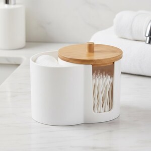 May include: A white, two-compartment bathroom organizer with a bamboo lid. One side holds cotton pads, the other holds cotton swabs. The lid has a small, cylindrical handle. The organizer is on a white countertop.