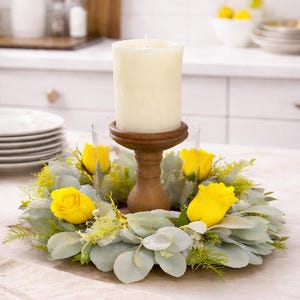 Puede incluir: Una vela de pilar color crema se encuentra sobre un soporte de madera dentro de un cilindro de vidrio, rodeada por una corona de hojas verde grisáceo y rosas amarillas. El arreglo está sobre una mesa con un paño de color neutro.