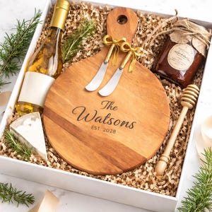 Puede incluir: Caja de regalo con una tabla de quesos de madera grabada con "The Watsons EST. 2028", dos cuchillos de queso, una botella de aceite de oliva, un tarro de mermelada, una cuña de queso y un cucharón de miel. La caja está llena de papel.