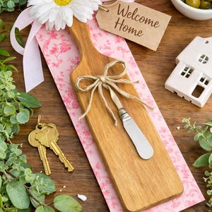 Puede incluir: Una tabla de queso de madera con un cuchillo plateado, atada con hilo, descansa sobre papel floral rosa. Una margarita blanca, una cinta rosa y una etiqueta que dice "Welcome Home" están adjuntas. Llaves doradas y una casita blanca.