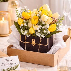 Puede incluir: Un arreglo floral de tulipanes amarillos y blancos y paniculata en un recipiente negro con forma de bolso con detalles de cadena dorada. El arreglo está en una caja marrón, con una caja de regalo y una tarjeta que dice "Home Sweet Home".