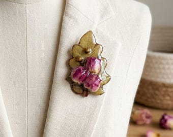 May include: A brooch featuring a preserved green oak leaf with a glossy finish, adorned with three dried magenta rosebuds and two small brown acorns. The botanical piece is attached to a light beige textured fabric garment, showcasing natural details.