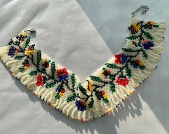 May include: A wide, curved collar necklace crafted from small white seed beads, featuring a repeating floral vine pattern in red, blue, green, and yellow. The edges are finished with a delicate fringe of white beads and a silver-toned metal clasp.