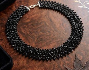 May include: A circular choker necklace constructed from small, uniform black beads arranged in a dense, interconnected lattice pattern. The necklace features a silver-toned metal lobster claw clasp closure at the back to secure the piece around the neck.