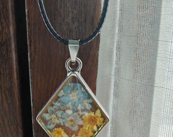 May include: A diamond-shaped pendant necklace featuring a clear resin setting filled with pressed light blue and yellow flowers. The pendant is attached to a simple black cord, creating a contrast between the floral elements and the dark necklace band.