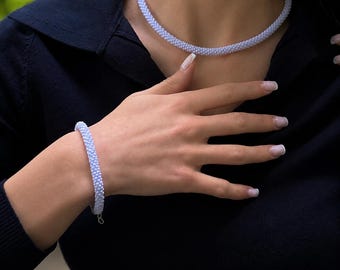 Może przedstawiać: Jasnoniebieski naszyjnik z koralików i pasująca do niego bransoletka. Obie ozdoby wykonane są z małych, jednolitych, okrągłych koralików ułożonych w gęsty, cylindryczny wzór, tworząc teksturowany, przypominający sznur wygląd na skórze.