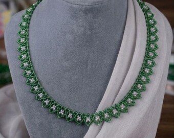 May include: A beaded necklace featuring a repeating pattern of small, dark green seed beads forming pointed, leaf-like shapes, each centered with a tiny, white, round bead. The necklace is arranged in a circular shape with a consistent design.