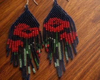May include: A pair of dangle earrings crafted from small beads in a triangular shape. The design features a dark blue background with a red floral pattern at the top and green beaded fringe hanging from the bottom, attached to silver-toned hooks.