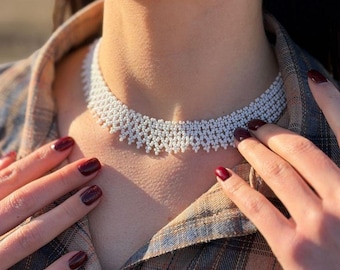 Może przedstawiać: Biały naszyjnik typu choker wykonany z koralików, o gęstym, misternym splocie z wykończeniem w kształcie łuków na dolnej krawędzi. Naszyjnik składa się z wielu drobnych, jednolitych białych koralików ułożonych w zwartą strukturę przylegającą do szyi.