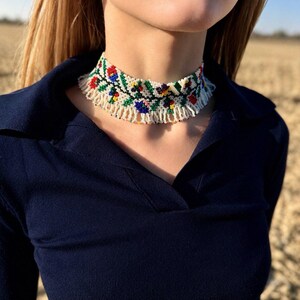 Może przedstawiać: Naszyjnik typu choker wykonany z koralik&oacute;w na białym tle z wielokolorowym wzorem kwiatowym w kolorach czerwonym, niebieskim, zielonym i ż&oacute;łtym. Naszyjnik posiada frędzle z białych koralik&oacute;w zwisające z dolnej części gł&oacute;wnej opaski.