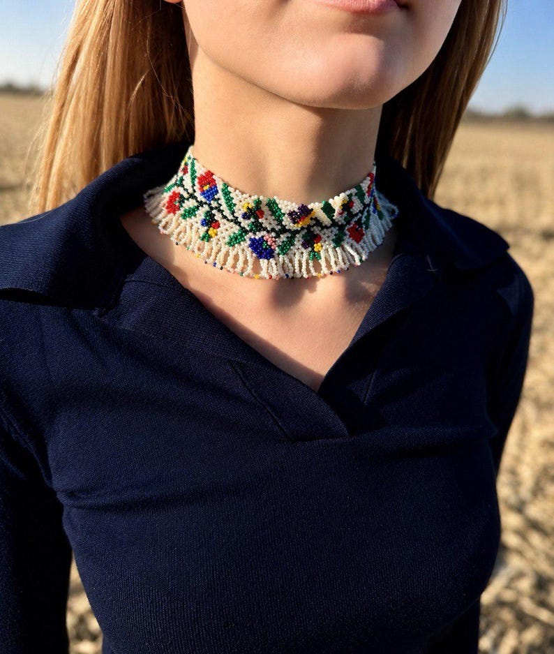 Może przedstawiać: Naszyjnik typu choker wykonany z koralik&oacute;w na białym tle z wielokolorowym wzorem kwiatowym w kolorach czerwonym, niebieskim, zielonym i ż&oacute;łtym. Naszyjnik posiada frędzle z białych koralik&oacute;w zwisające z dolnej części gł&oacute;wnej opaski.