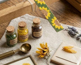 May include: A wooden bead loom with a partially woven sunflower pattern strip, three small glass jars containing olive green, sunflower yellow, and espresso center beads, a brass scoop, a sunflower print in a frame, and a spool of twine.