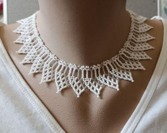 May include: A white beaded necklace featuring a repeating pointed, leaf-like pattern constructed from small, uniform white beads. The design creates a structured, collar-style shape that sits closely around the neck, showcasing intricate beadwork.
