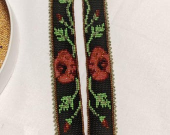 May include: A long, narrow beaded necklace featuring a black background with a floral pattern of red poppies and light green leaves. The edges of the necklace are finished with a border of small, metallic gold beads, creating a structured design.